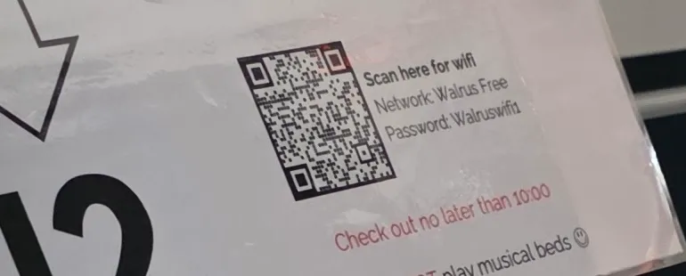 Crop of a laminated card on a hostel bunk bed: a QR code next to 'Scan here for wifi — Network: Walrus Free — Password: Walruswifi1', above 'Check out no later than 10:00'