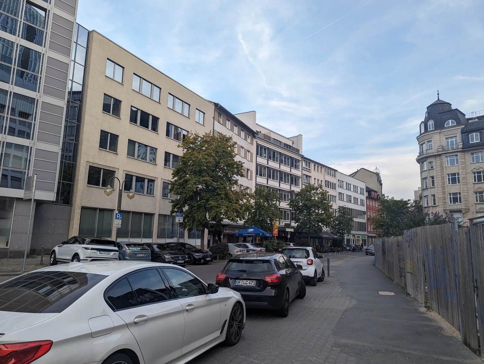 The challenge photo: a street of post-war office buildings with trees, a row of parked cars on the left, a wooden hoarding with graffiti on the right, and a sandstone corner building with a slate dome at the far right
