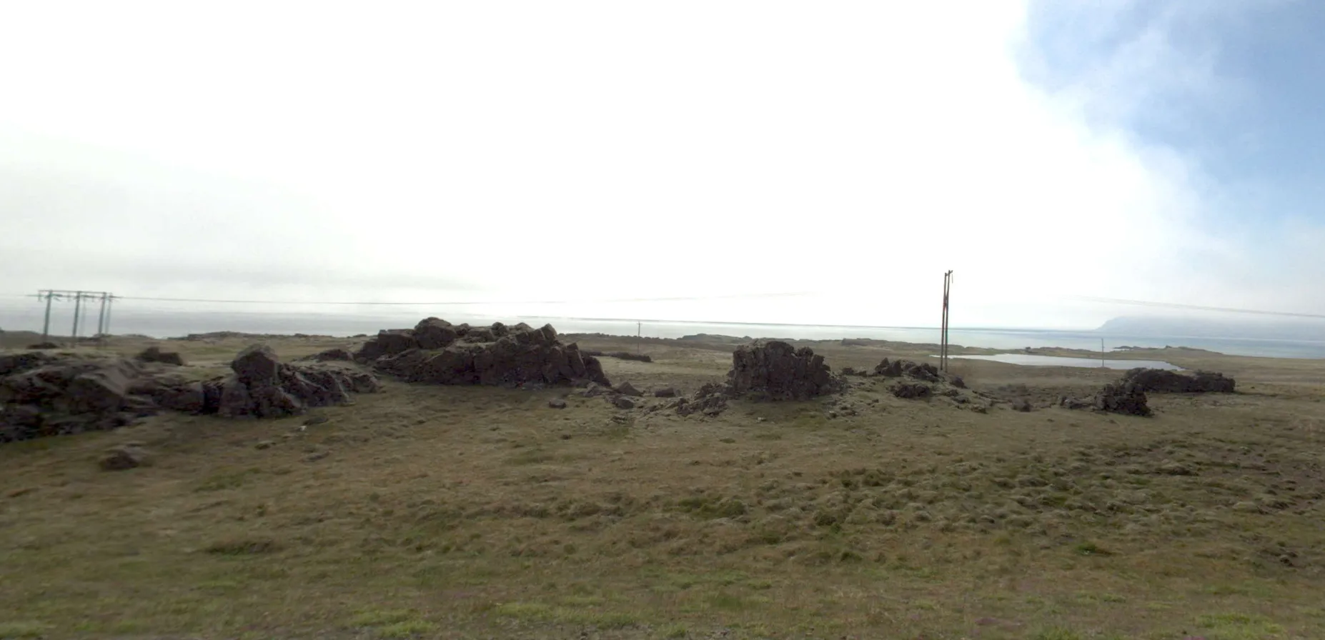 The challenge image: a treeless coastal plain under a flat white sky, dark basalt outcrops across the middle distance, a shallow lagoon and open water to the right, a low pole line at the left edge and a taller two-legged wooden pole right of centre