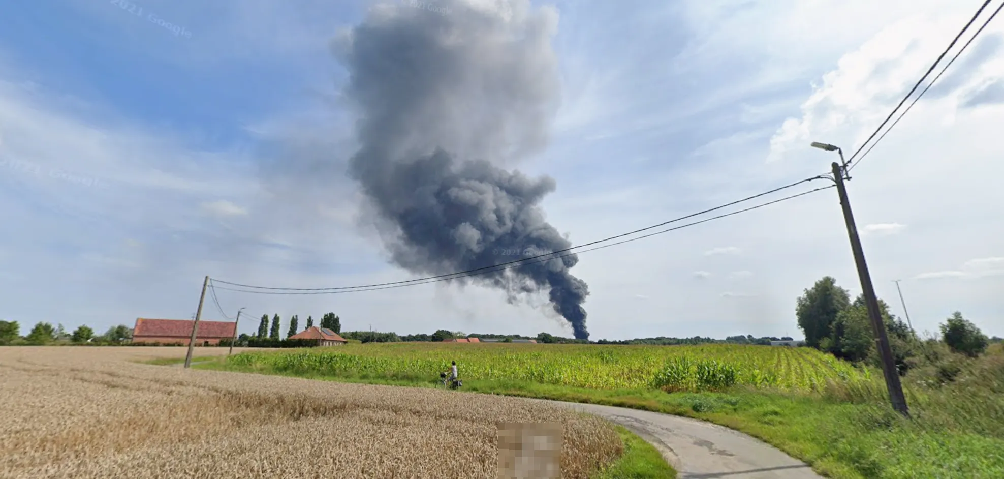 The challenge image: a narrow farm road curving through a ripe wheat field on the left and a maize field on the right, a cyclist on the road, a red-brick farmhouse and a row of poplars on the horizon, concrete poles carrying overhead wires and a street lamp, and a tall column of black smoke rising behind the fields