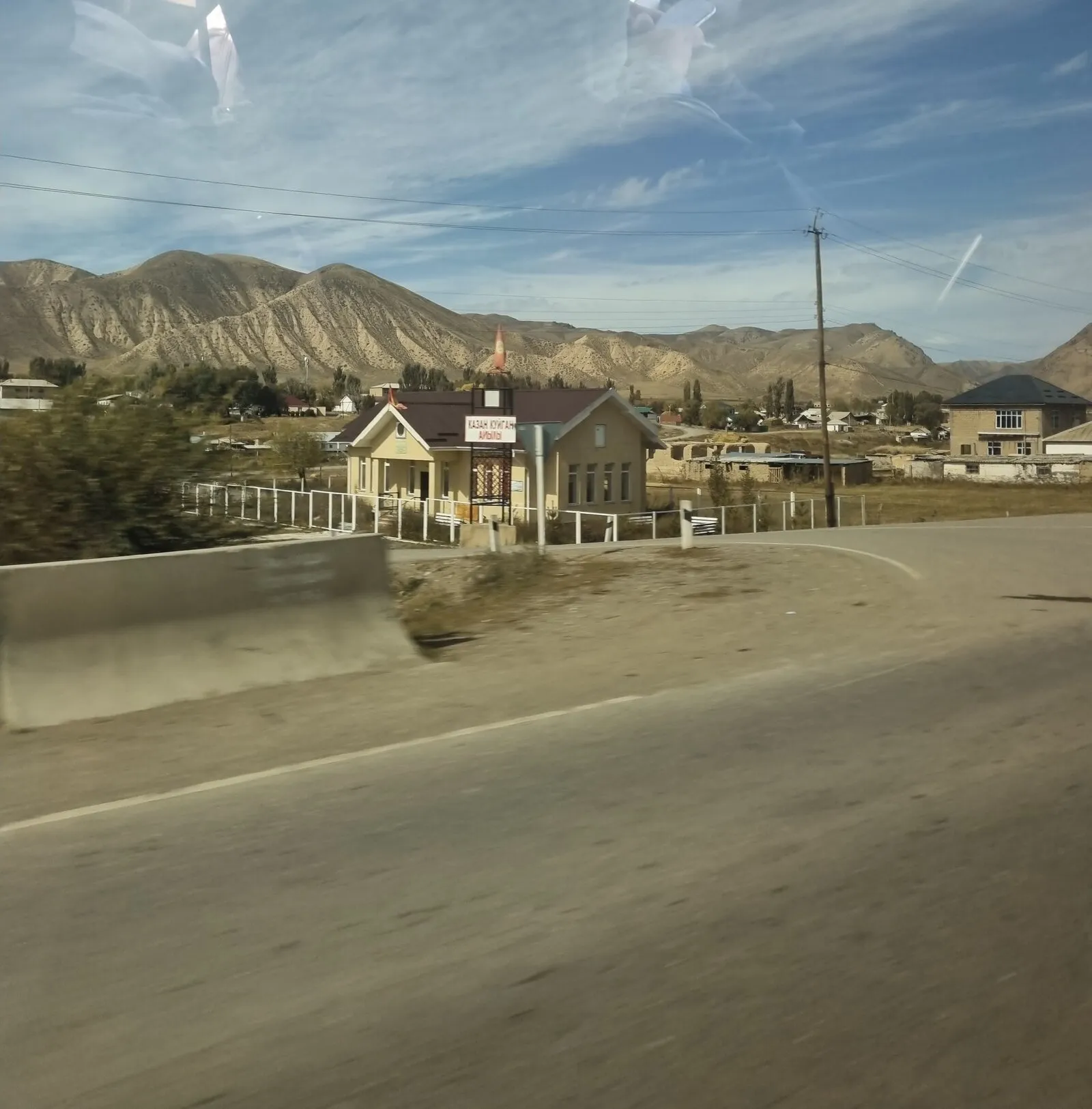 The challenge photo, taken through a car window: window reflections across the sky, a pale yellow single-storey building with a brown roof and a small flag behind a white fence, a sign on a black frame in front of it, eroded tan mountains behind, and a blurred road with a concrete barrier in the foreground