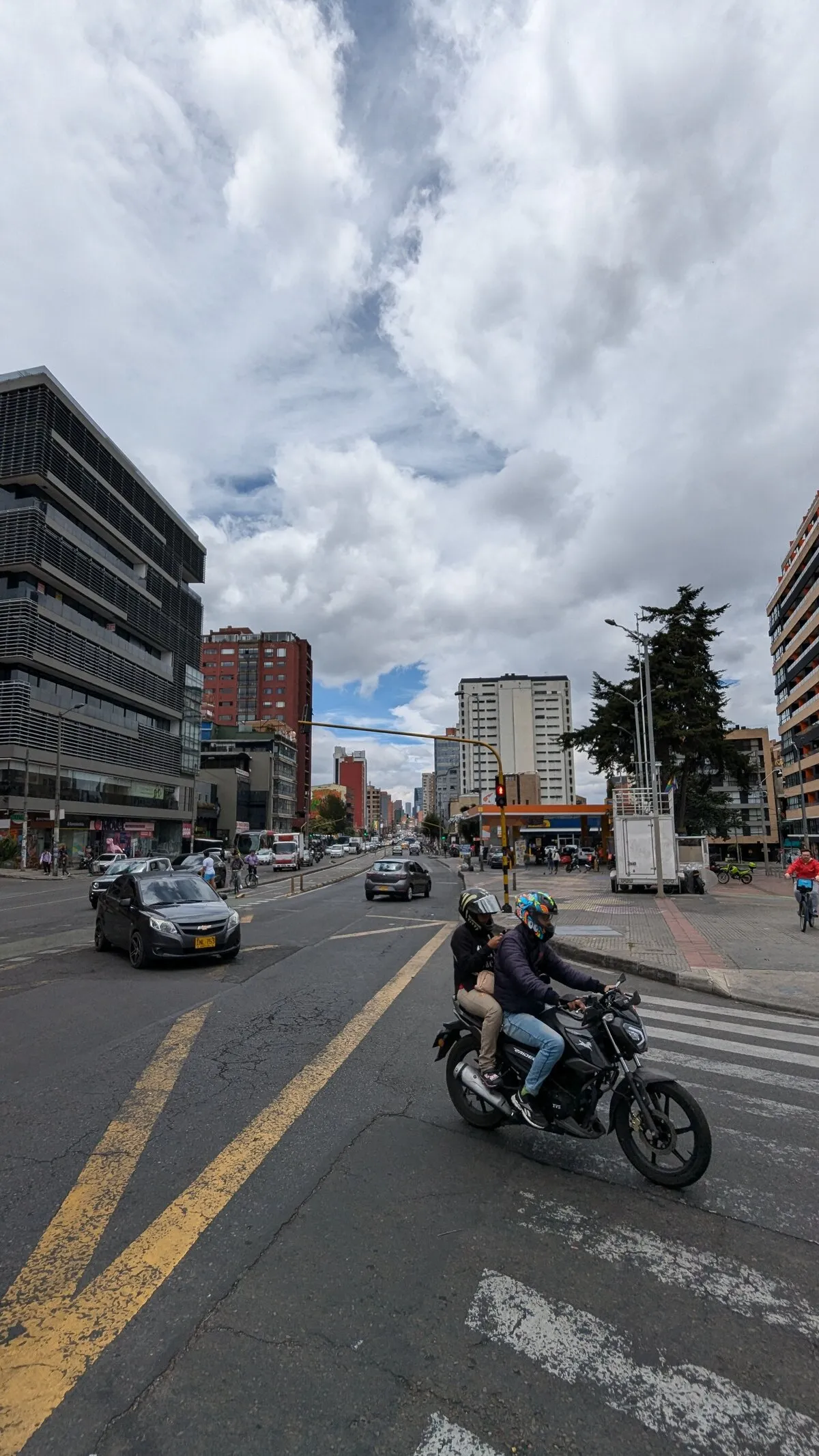 The challenge photo, portrait format: a wide street under a cloudy sky, a dark office block with horizontal louvres on the left, high-rises and a yellow traffic light arm in the middle, a plaza with a tall conifer and a white trailer on the right, and in the foreground two people on a black motorbike crossing a zebra crossing