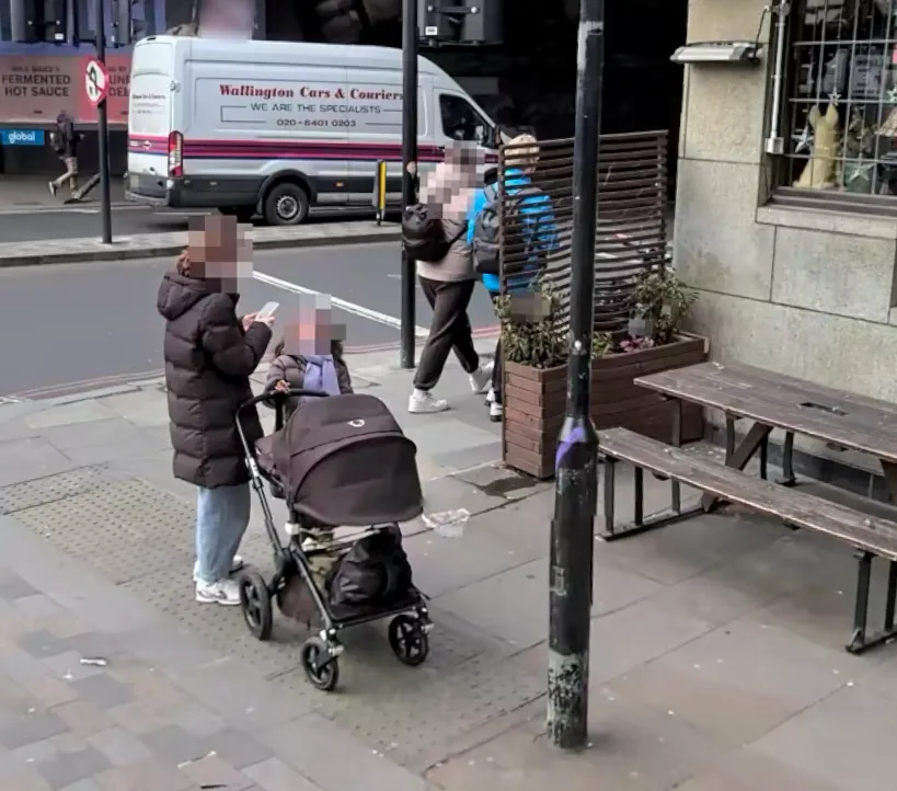 The challenge photo: a woman with a pram on a wide paved London pavement, a white Wallington Cars & Couriers van across the road, a red 'Fermented Hot Sauce' advert behind it, and a stone-fronted pub with picnic benches to the right, all under a dark overhead structure