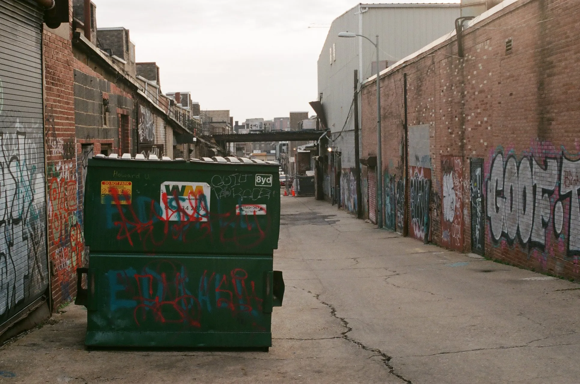 The challenge photo: a graffiti-covered green Waste Management dumpster in a narrow alley, brick buildings on the left, a brick wall with large graffiti on the right, a steel bridge crossing the alley in the mid-distance and a city skyline at the far end