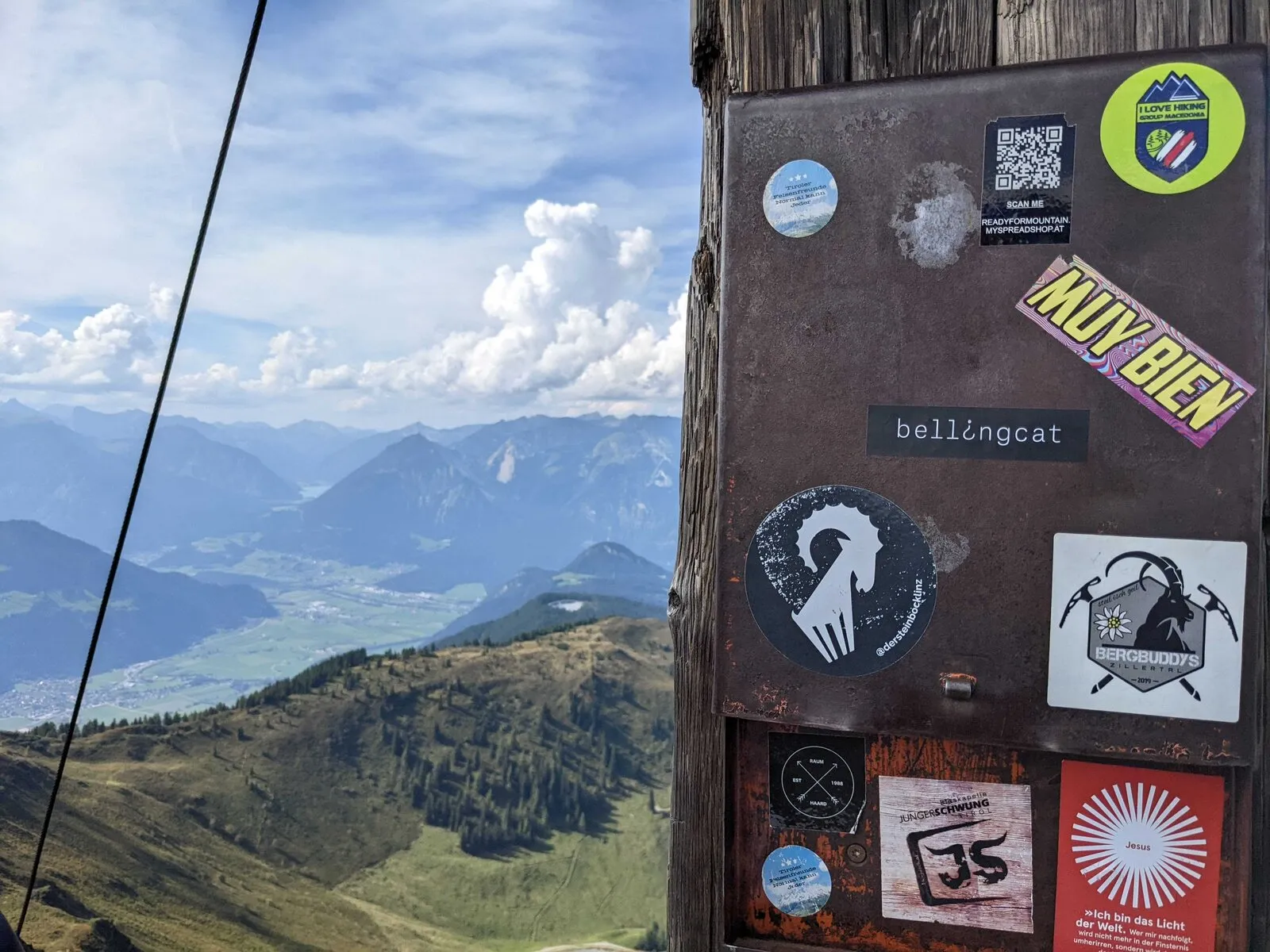 The challenge photo: on the left a steel cable runs diagonally across a view over a wide green valley with mountains behind it; on the right a weathered wooden post with a rusty metal box covered in stickers, among them a black bellingcat strip, a QR code sticker, a green I LOVE HIKING circle, a MUY BIEN strip, a white ibex logo and a Bergbuddys Zillertal sticker