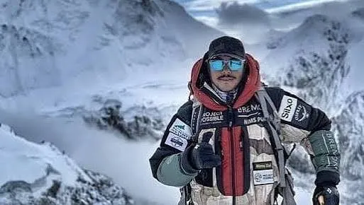 The challenge image: a mountaineer in a Project Possible down suit giving a thumbs-up on a snowed-in ridge, glacier and peaks behind him