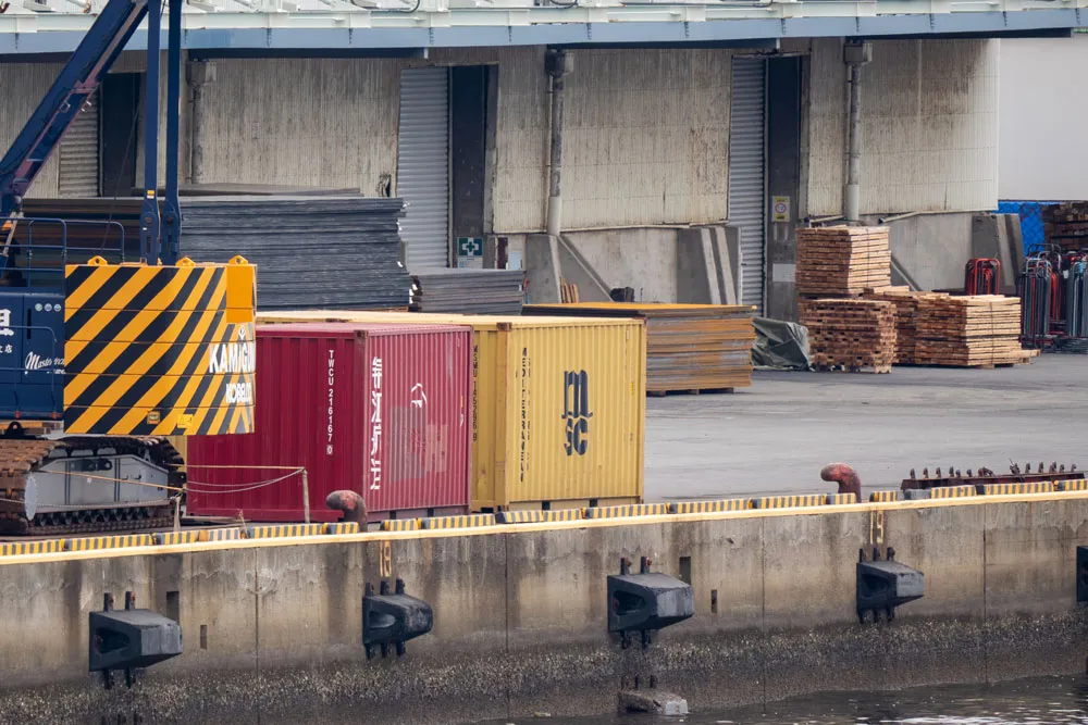 Quayside with a yellow MSC container, a red container beside it, and a Kamigumi gantry crane in front of a warehouse