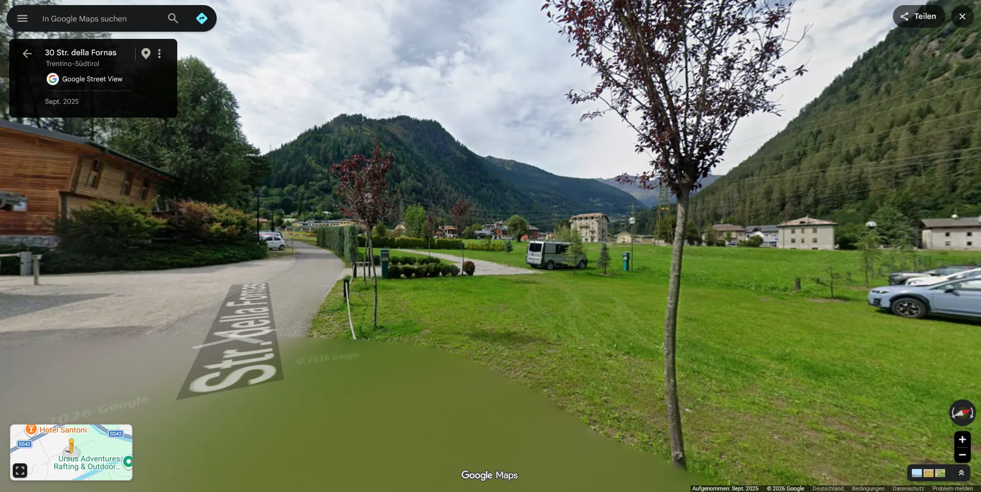 Google Street View on Str. della Fornas in Ossana: gravel lot and hedge-lined cycle path in the foreground, the valley falling away toward an identical mountain profile