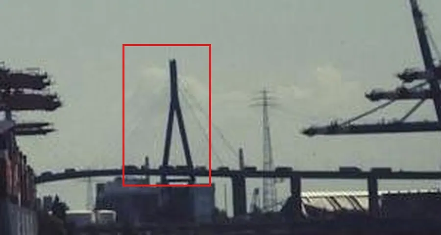 An enlarged crop of the horizon: a long elevated road bridge with a single tall A-shaped pylon, with stay cables fanning out, in a red box, a lattice electricity pylon to its right and crane booms at the edges