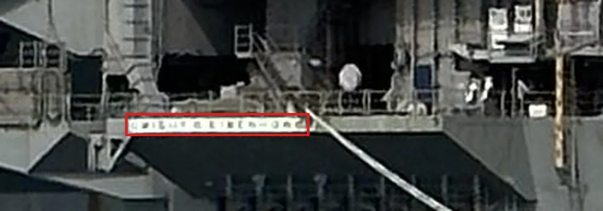 An enlarged crop of the carrier stern in the middle of the image: a long white nameplate with dark capital letters below the hangar deck opening, in a red box