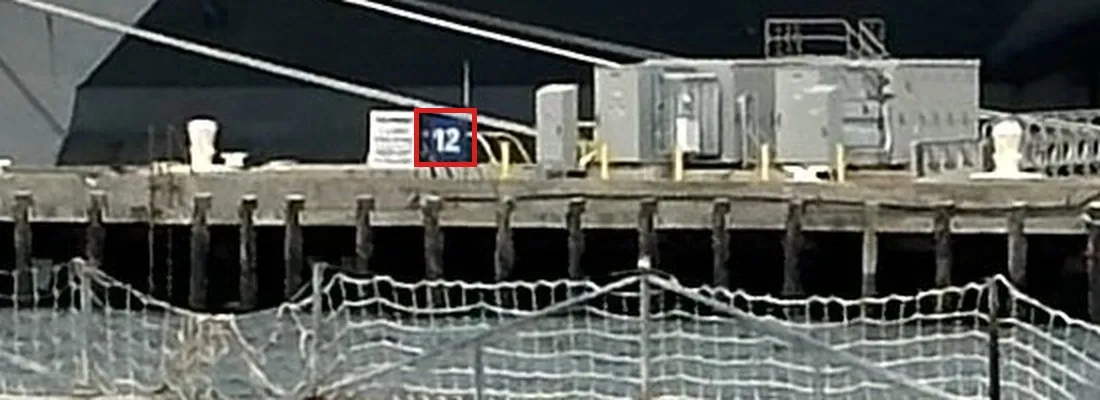 An enlarged crop of the pier in front of Ike: a white notice board and next to it a small blue sign with the white number 12 in a red box, on a concrete pier deck above wooden piles, with the security netting in the foreground