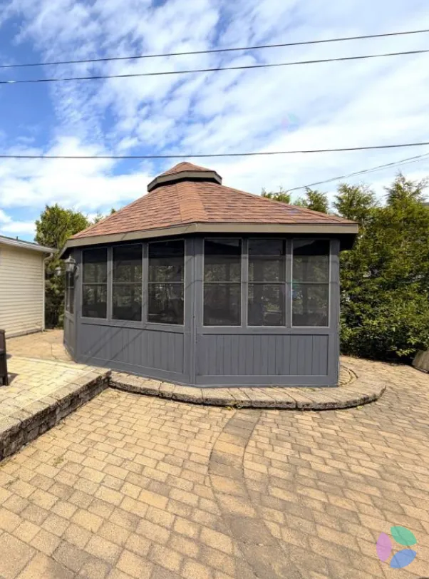A grey screened octagonal gazebo with a shingled roof and cupola, standing on a paver patio beside a beige house, hedge behind, overhead power lines.