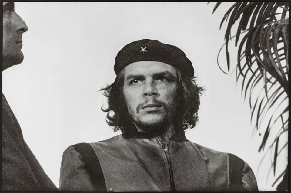 The uncropped frame of Guerrillero Heroico: Che Guevara in a beret, with a man's profile at the left edge and palm fronds at the right — the parts the famous crop removes