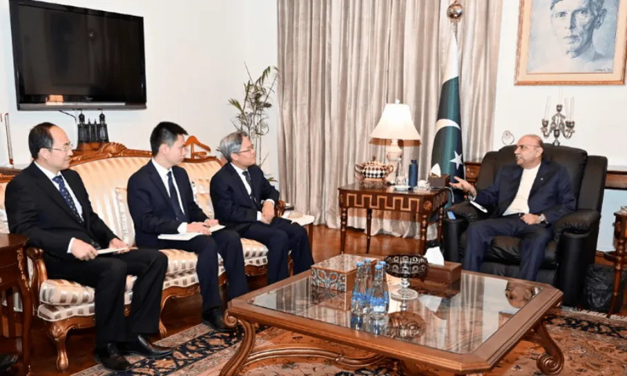 Uncaptioned wire photo: four visitors seated on one side, one host on the other, national flag and a founder's portrait in the background.