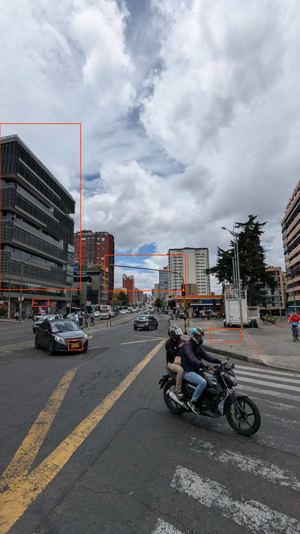 The challenge photo with red boxes: the dark office block with louvres on the left, a shop sign at its base, the yellow licence plate of a car, the traffic light arm with the towers behind it, and a paved patch on the plaza