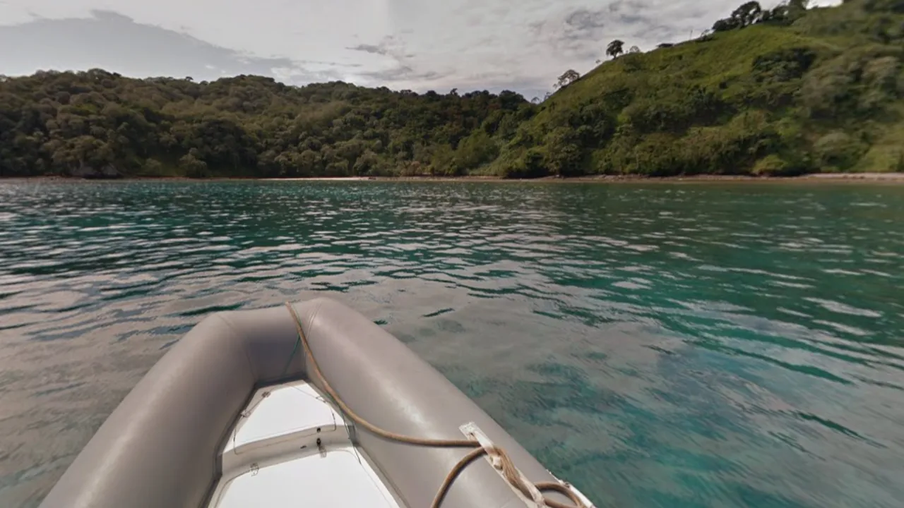 Street View frame taken from the bow of a grey inflatable boat, its rope and cleats in the foreground, over clear turquoise water towards a steep shoreline of dense green forest with a narrow beach at its foot