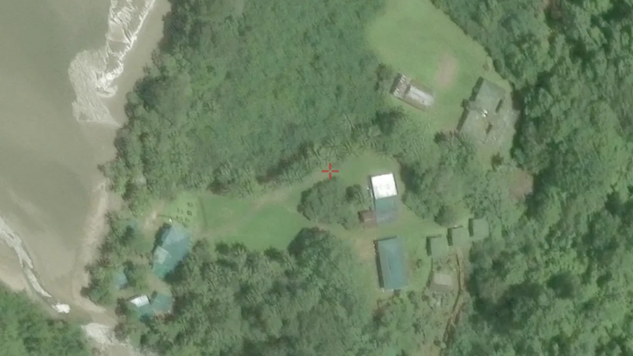 Satellite crop with a small red Google Earth crosshair near the centre: a clearing in dense rainforest on a headland, two long light-roofed sheds and several smaller teal-roofed buildings around a mown lawn, footpaths running down to a bay whose water is brown with river sediment