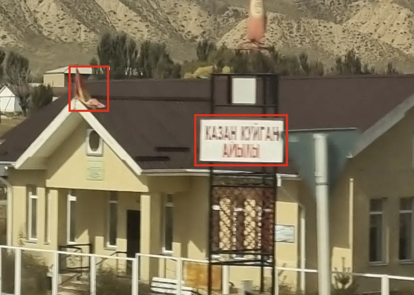 An enlarged crop of the building: a red box around the sign reading КАЗАН КҮЙГАН АЙЫЛЫ in red letters, half blurred, and a second box around a red flag with a yellow centre on the roof ridge