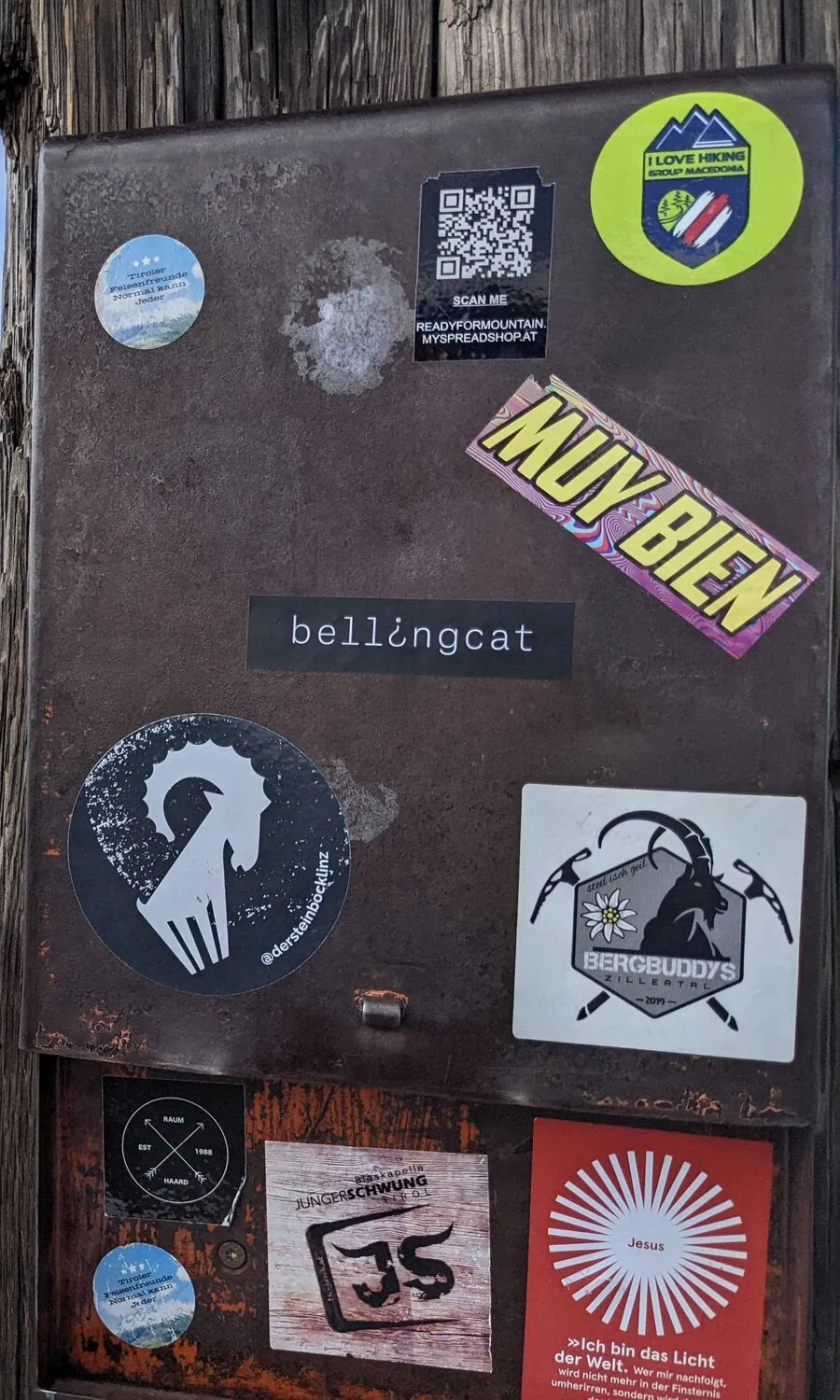 A close crop of the sticker box: a small round Tiroler Felsenfreunde sticker, a QR code labelled SCAN ME READYFORMOUNTAIN.MYSPREADSHOP.AT, a green I LOVE HIKING GROUP MACEDONIA circle, a MUY BIEN strip, the bellingcat strip, a round ibex sticker signed @dersteinbocklinz, a Bergbuddys Zillertal square, and below a Jungerschwung sticker and a red Jesus sticker