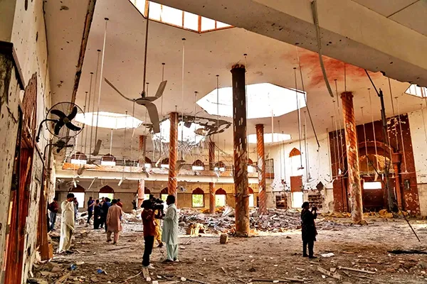 Interior of a large damaged prayer hall: several blown-open dome apertures in the ceiling, patterned columns, ceiling fans, rubble across the floor, and press photographers standing among it.