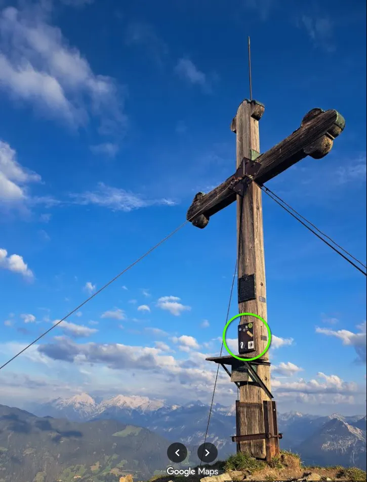 A Google Maps user photo of the Wiedersberger Horn, September 2022: a tall wooden summit cross against a blue sky, held by steel guy wires, with a green circle around a small dark box with stickers on the post just above a wooden shelf