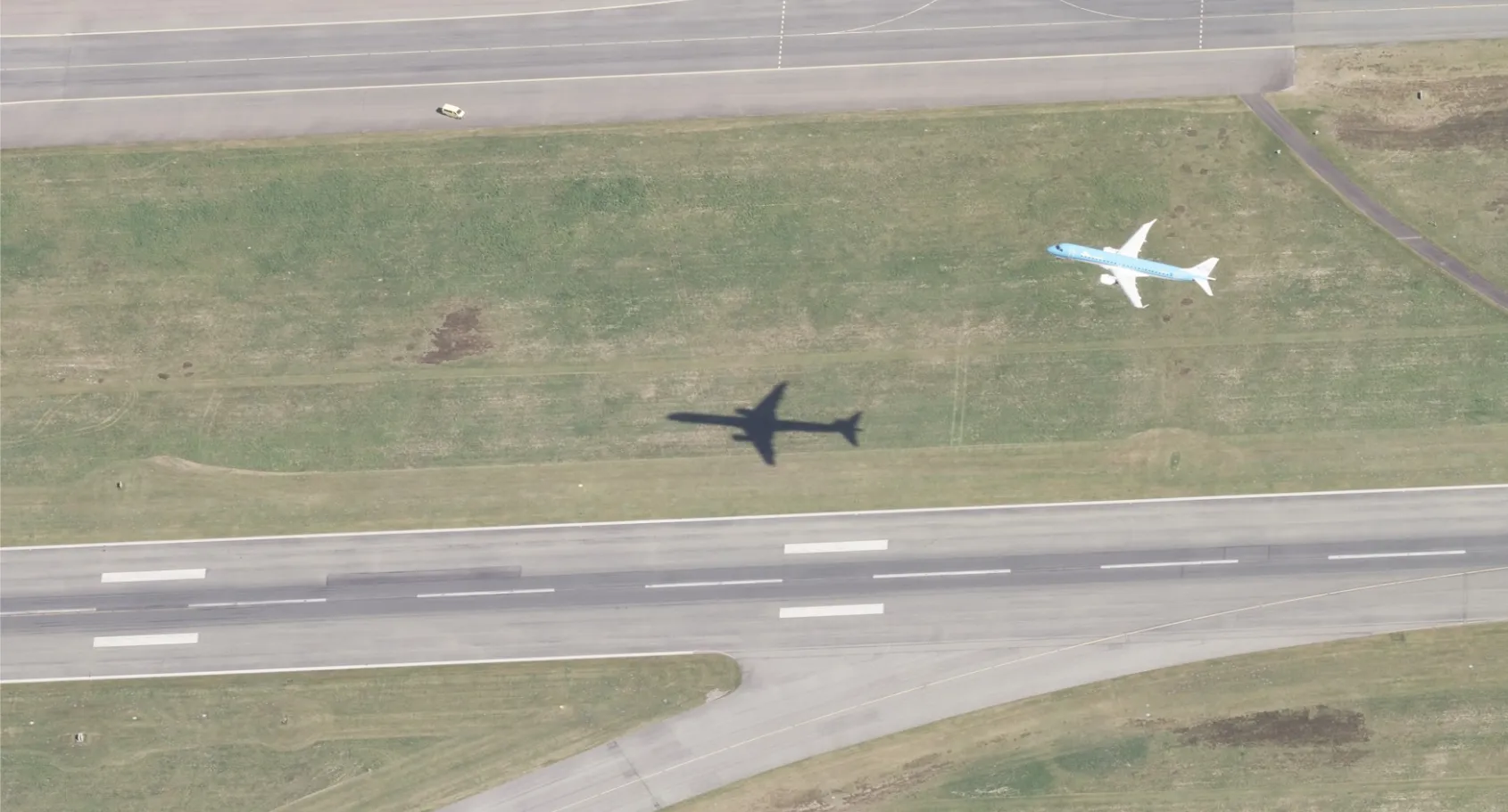Aerial view of a light blue airliner climbing over a grass strip between a runway and a taxiway
